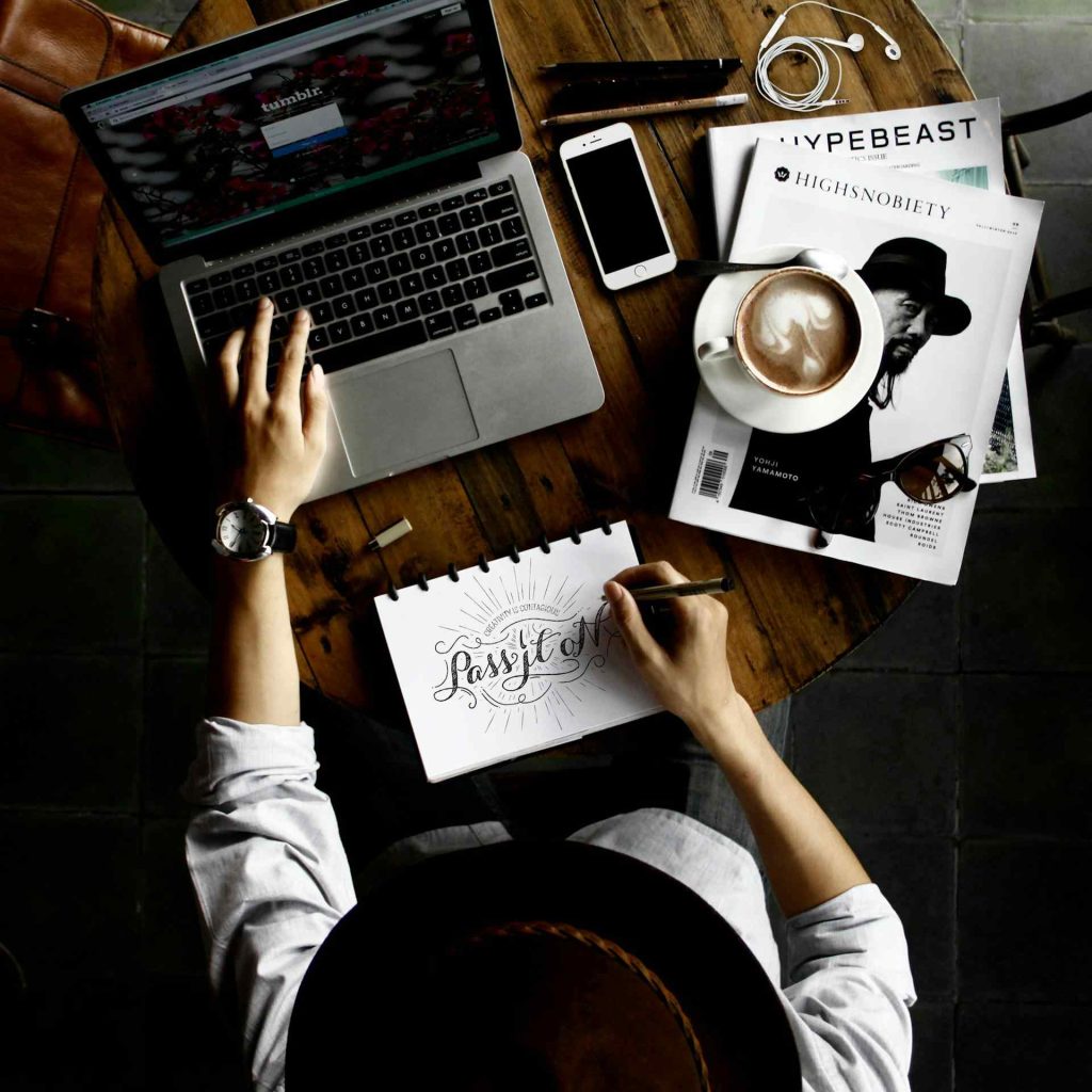 Overhead View Of a Person In His Workspace With Laptop, Sketchbook, Coffee, And Phone On a Wooden Table creating & brain storming Graphic Design Solutions with 27 DiGi