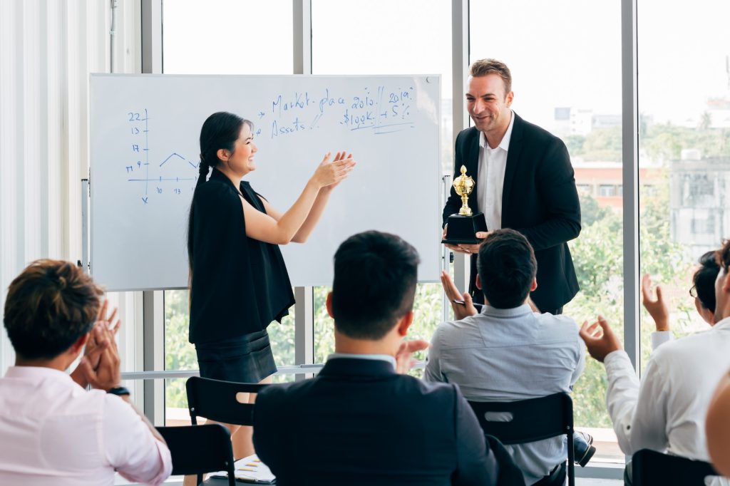 Woman Presenting To Audience and talking about us 27 digi With Man Holding Trophy, Whiteboard With Graphs In Background.