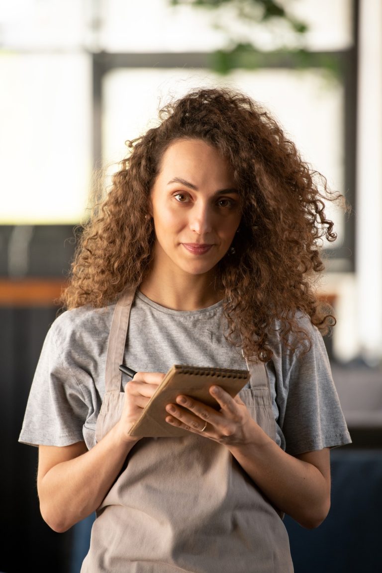 A Woman With Curly Hair In An Apron Writing In a Notebook about the FAQs of 27 DiGi