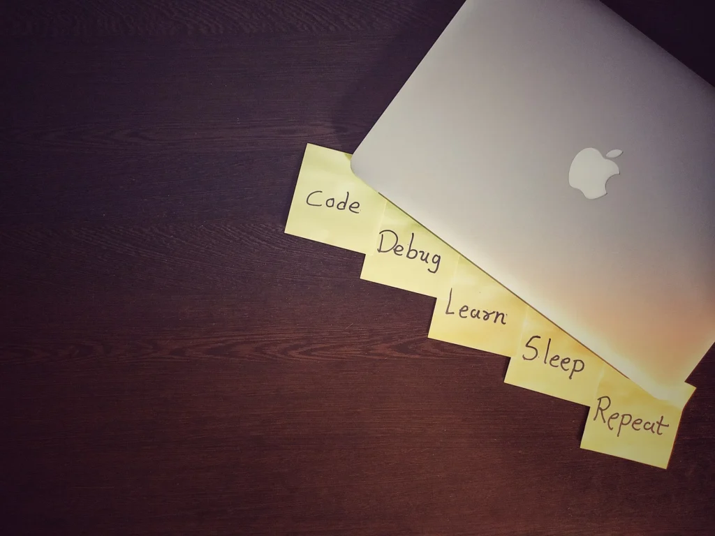 A MacBook rests on a wooden surface, surrounded by sticky notes labeled: "Code, Debug, Learn, Sleep, Repeat."