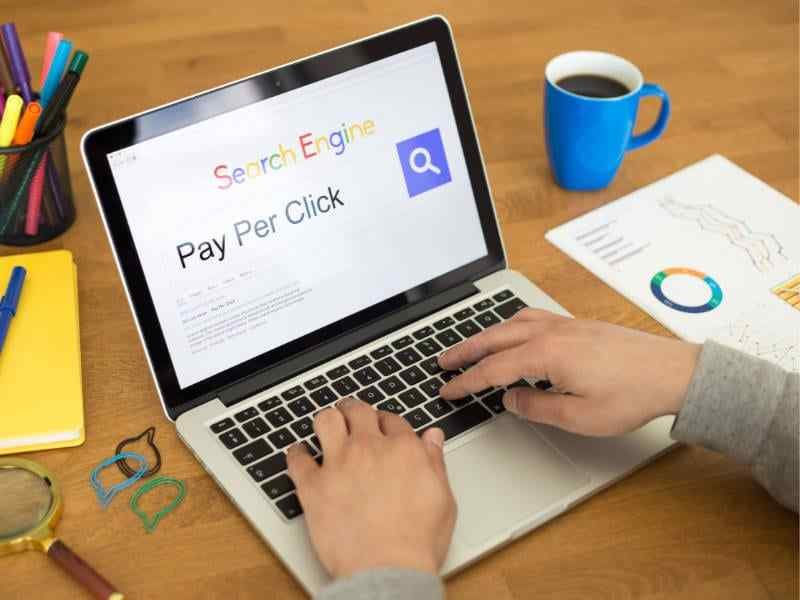 Person Using Laptop Showing "Pay Per Click" On Screen, With Coffee And Analytical Charts On Desk.